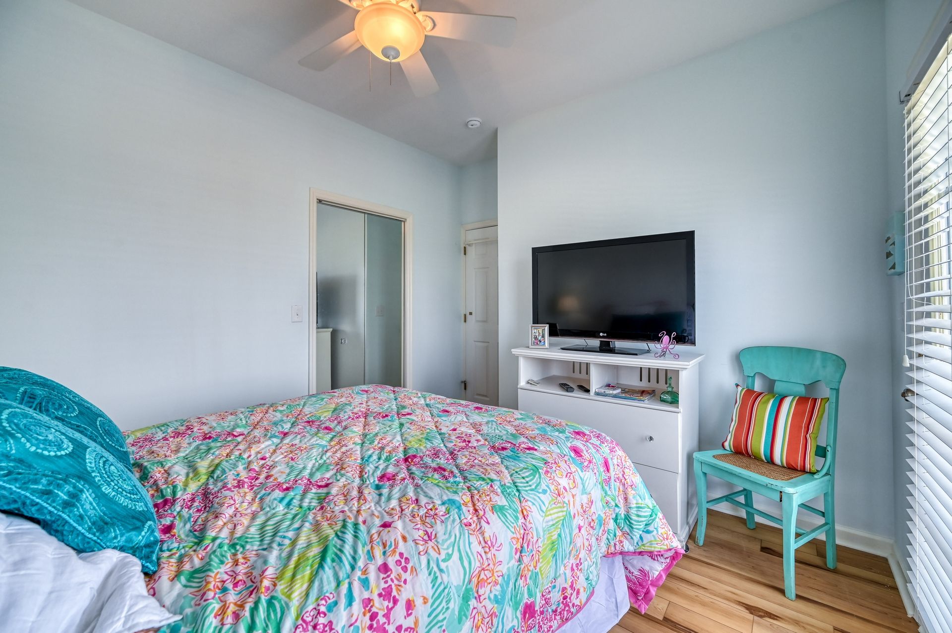 A bedroom with a bed, a TV on a stand, a chair, and a window with blinds.