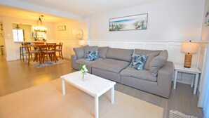 A living room with a grey sofa, a white coffee table, and a dining area with wooden chairs and a chandelier. - The Chateau 101B-Baywater 9thth St, W/D, AC, Free WIFI (Ocean City)