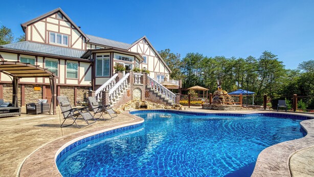 A swimming pool with a stone deck, a covered patio, and a two-story house with a balcony. - Fabulous Private Pool with Waterfall! Tudor Rose Lodge (Sevierville)
