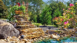 A garden with a water feature, including a stone waterfall and a pond surrounded by rocks and plants. - Fabulous Private Pool with Waterfall! Tudor Rose Lodge (Sevierville)
