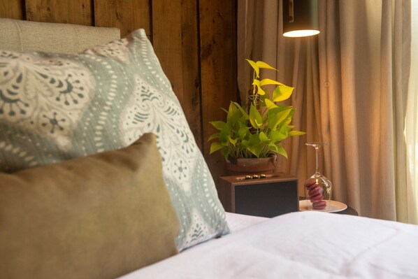 A bedroom with a wooden headboard, patterned pillows, a bedside table with a plant, a wine glass, and a lamp.