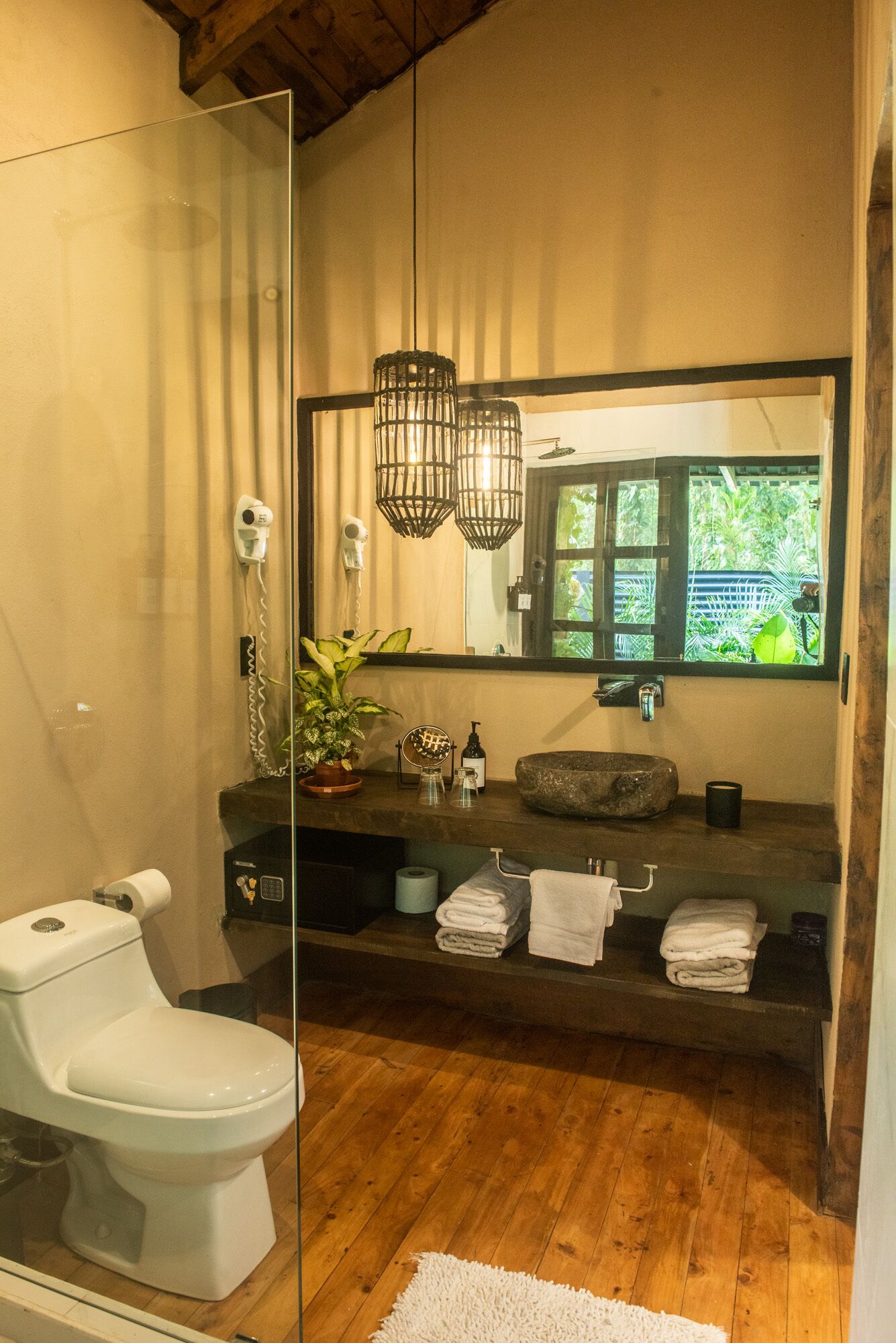 A modern bathroom with a glass shower enclosure, a white toilet, a wooden vanity with a large mirror, and a potted plant.