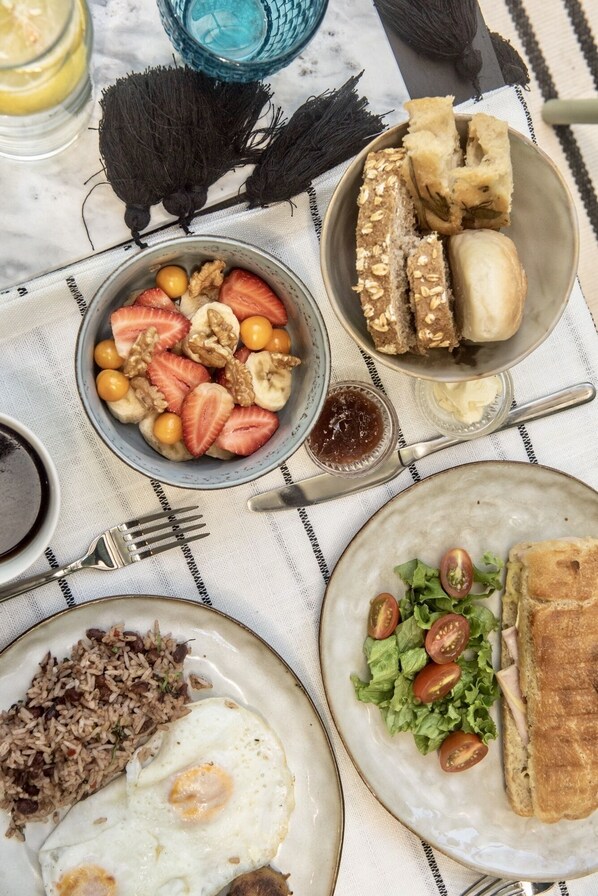 Una mesa con diversos alimentos para el desayuno, incluyendo un tazón de fruta, pan, un sándwich, arroz frito y un plato con huevos.