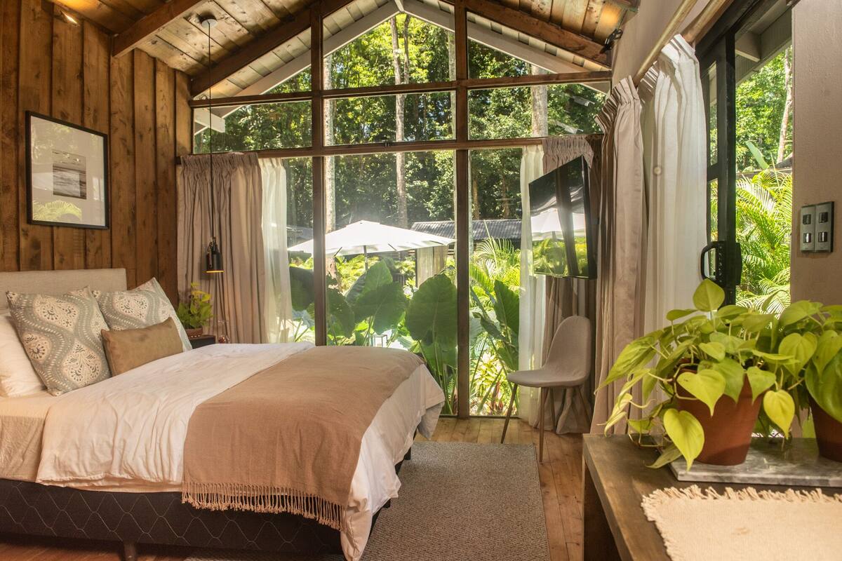Un dormitorio acogedor con un ventanal grande, una cama con almohadones estampados y una planta en maceta.