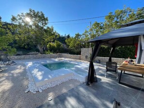 A swimming pool with a stone wall, a covered seating area, and a patio with a table and chairs.