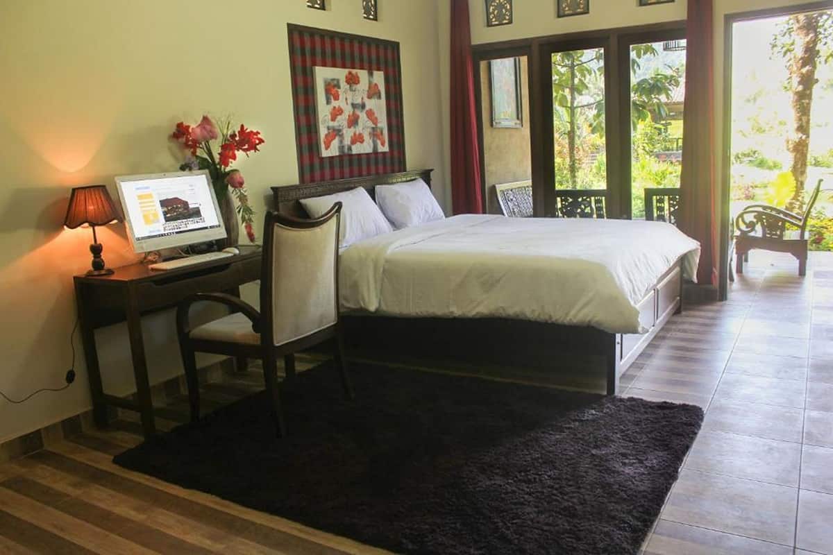 A bedroom with a bed, a desk with a computer, a chair, and a window with a view of greenery.