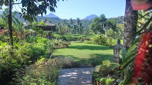 A lush garden pathway leading to a traditional structure with a tiled roof, surrounded by tropical vegetation and a mountain in the background. - Amartya Puri Green Cottages (Munduk)