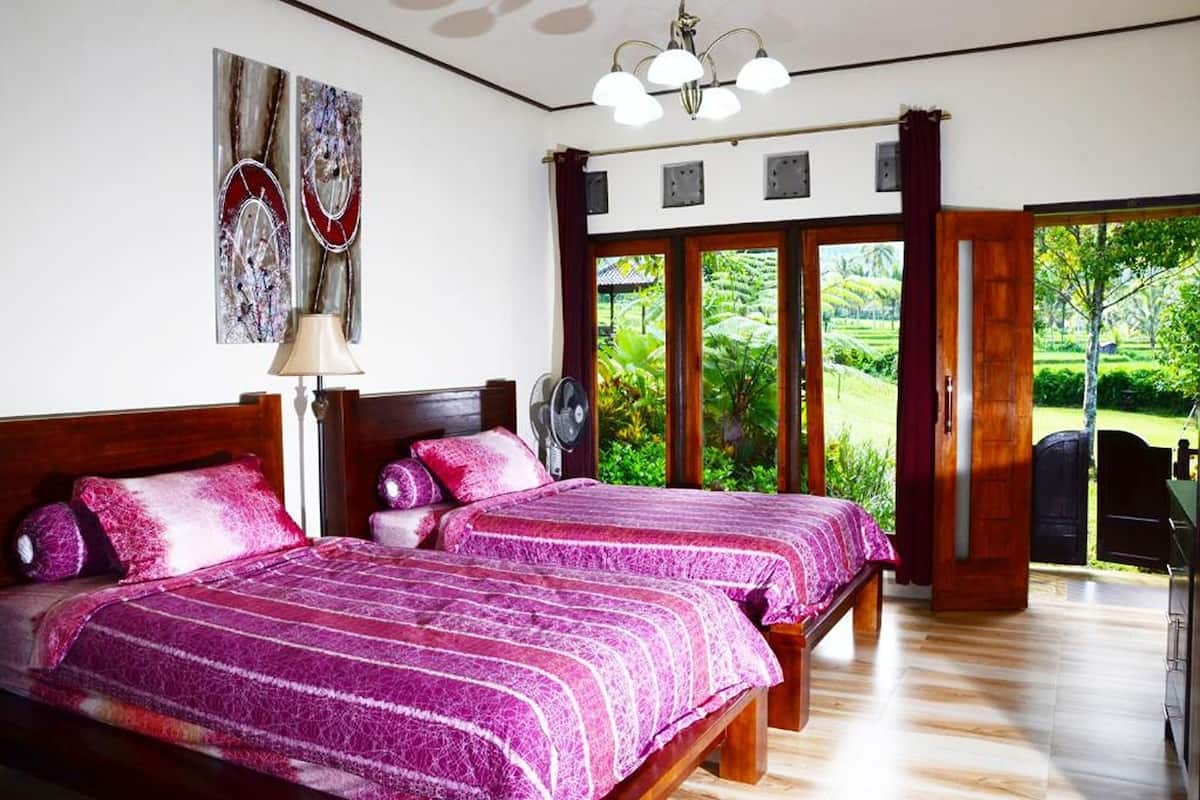 A bedroom with two beds, a wooden headboard, a ceiling fan, and a view of a lush garden through a sliding door.