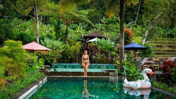 A woman standing by a pool with a swan float, surrounded by lush greenery and a small pavilion. - Amartya Puri Green Cottages (Munduk)