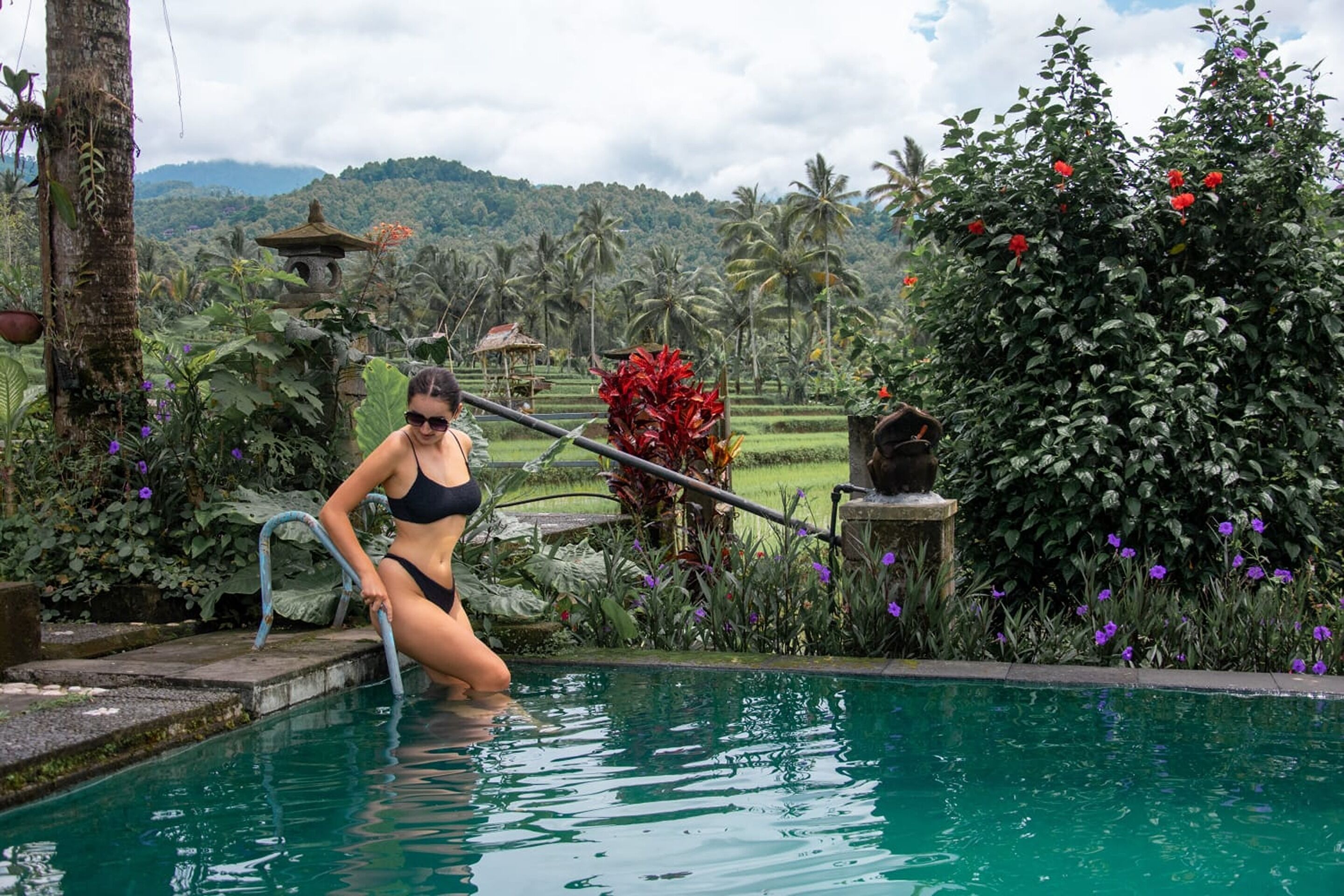 A woman in a black bikini sitting by a pool with a lush garden and a temple in the background.
