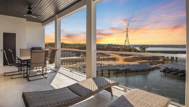 A balcony with wicker furniture, a dining area, and a view of a sunset over a river with a bridge and power lines. - Brand New Complex, Central Location, Amazing Views (Osage Beach)