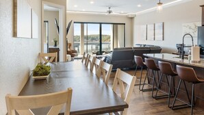 A spacious dining area with a long wooden table and multiple chairs, a kitchen area with modern appliances, and large windows offering a view of the outdoors. - Brand New Complex, Central Location, Amazing Views (Osage Beach)