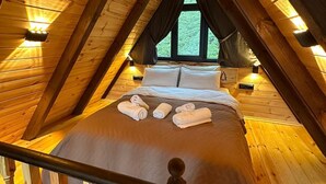Een gezellig slaapkamer op zolder met een schuine kap, houten balken, een bed met kussens en opgerolde handdoeken, en een raam met uitzicht op groen buiten.