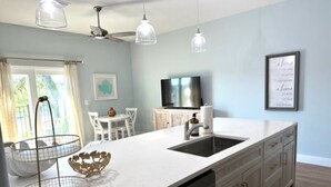 Eine moderne Küche mit weißer Arbeitsplatte, integriertem Geschirrspüler und einer großen Kochinsel mit Spüle. Es gibt einen Essbereich mit weißem Tisch und Stühlen sowie einen an der Wand montierten Fernseher.