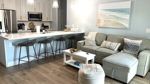 Eine moderne Küche mit Kochinsel, Barhocker, einem grauen Sofa und einem großen Gemälde zum Strandthema an der Wand.