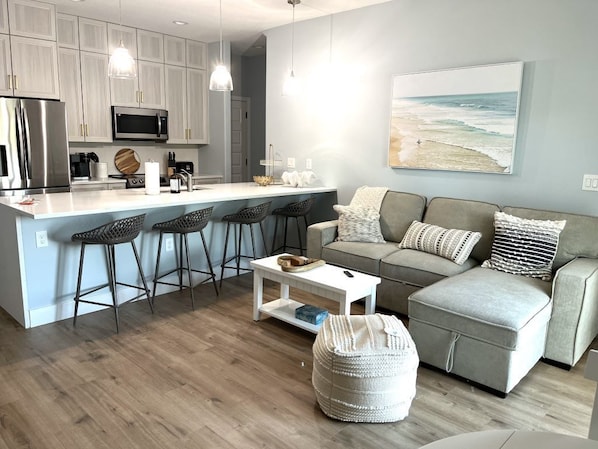 Una cocina moderna con una isla central, taburetes de bar, un sofá gris y un cuadro grande con temática marina.