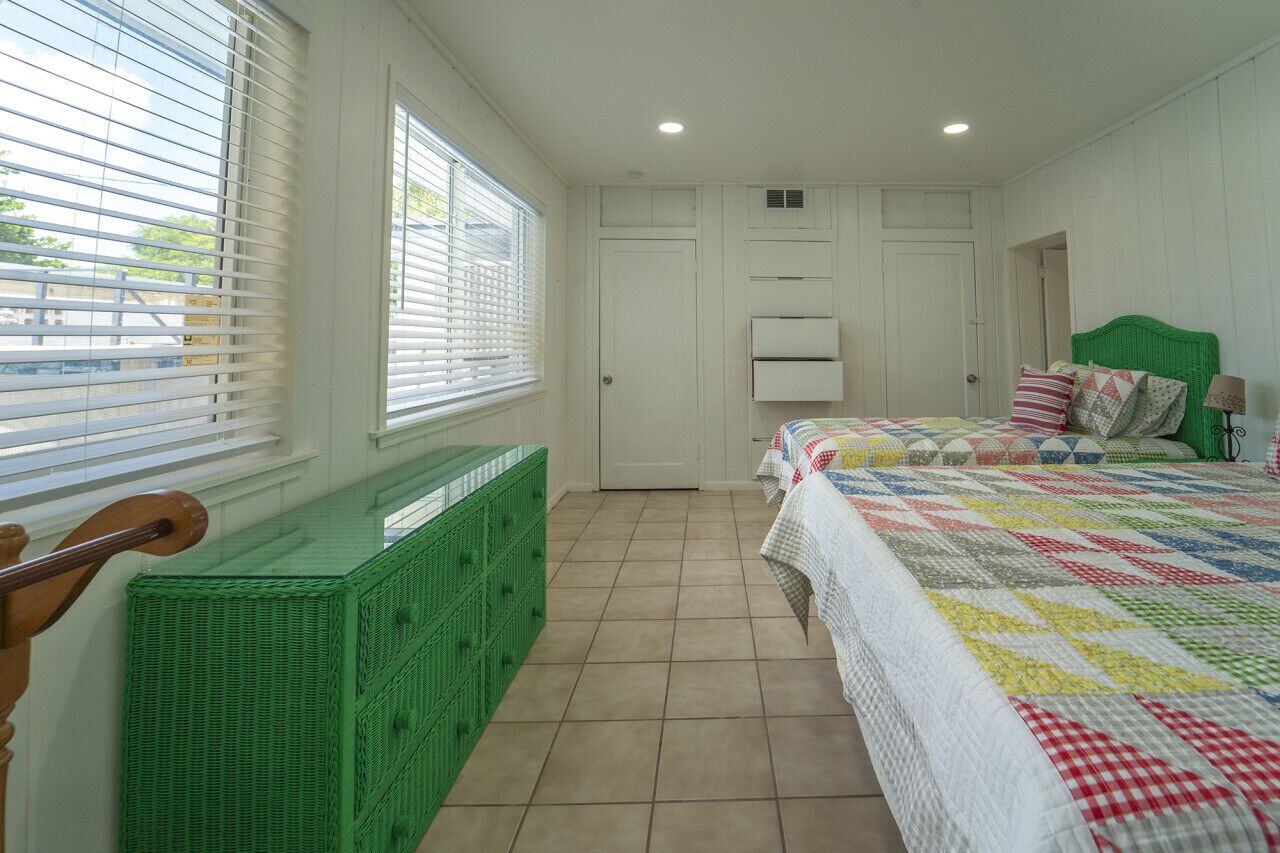 Een slaapkamer met een bed, een raam met zonwering, een groene rieten bankje en een witte deur.