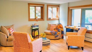 Una sala de estar acogedora con sillones estampados, un sofá y una mesita de centro. Hay ventanas con persianas, una pequeña mesita con libros y vistas al exterior.