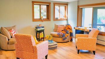 A cozy living room with patterned armchairs, a sofa, and a coffee table. There are windows with blinds, a small side table with books, and a view of the outdoors.