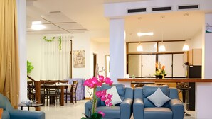 A modern living room with a blue sofa, a yellow ottoman, and a glass coffee table. There is a dining area with wooden furniture and a kitchen in the background. - Laciaville Hotel Deluxe Villa (Lapu Lapu)
