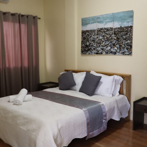 Een net opgemaakt bed met een wit-grijze deken, een houten hoofdbord en twee nachtkastjes met zwarte oppervlakken.