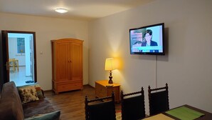 A living room with a television mounted on the wall, a wooden wardrobe, a wooden dining table with chairs, and a lamp on a side table.