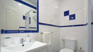 A bathroom with a white sink, mirror, and toilet, blue-tiled walls, and a blue-tiled shower area. - Tejares 16, Apartment 2 bedrooms Ground Floor (Sevilla)