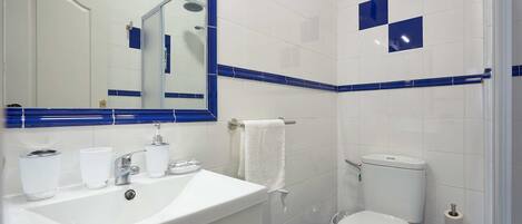 Un baño con lavamanos, espejo y retrete blancos, paredes y zona de ducha con azulejos azules.