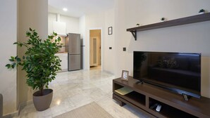 A modern living room with a flat-screen TV, a large potted plant, and a kitchen in the background. - Tejares 16, Apartment 2 bedrooms Ground Floor (Sevilla)