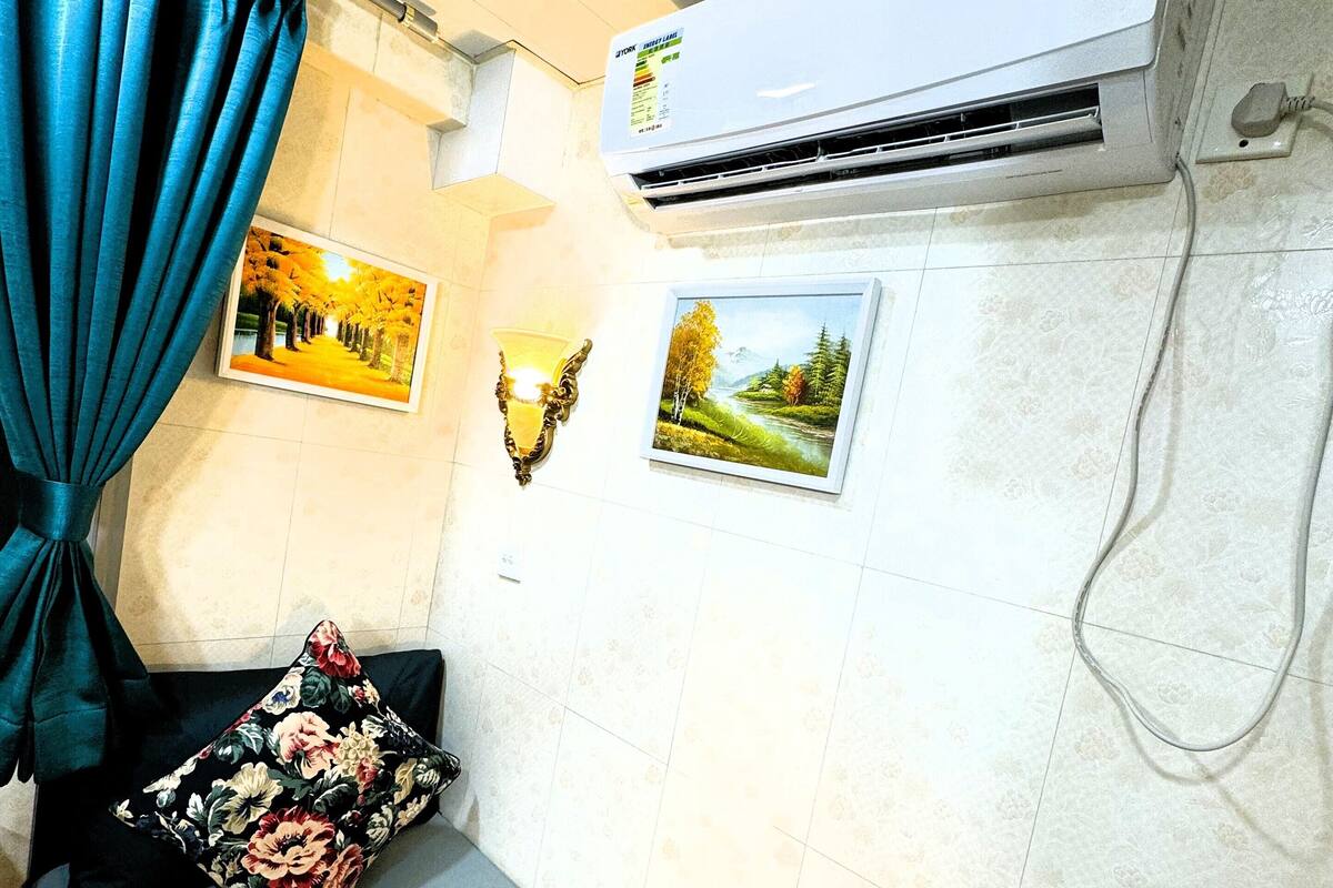 A room with a bed, a wall-mounted air conditioner, two framed pictures, a wall sconce, and a curtain.