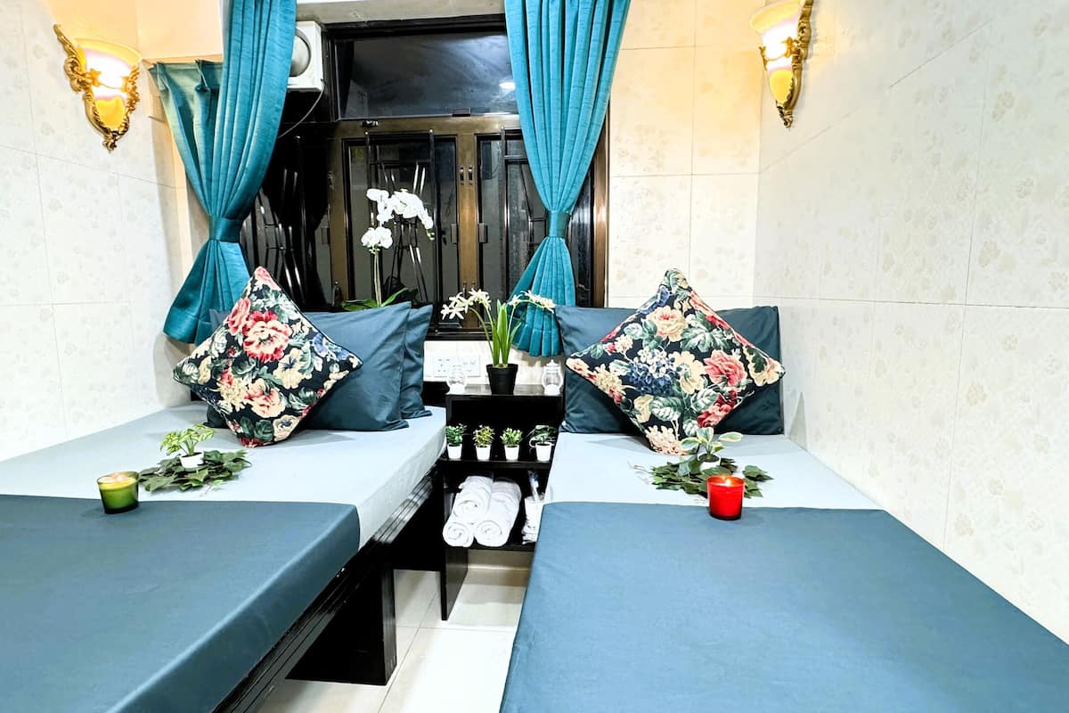 A room with two beds, each with a blue bedspread and floral pillows. There are potted plants on the beds and a small table with folded towels.