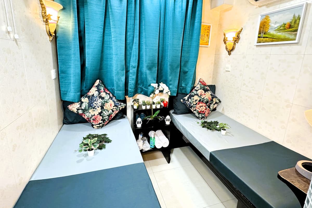 A room with two single beds, teal curtains, floral cushions, wall-mounted lights, a small shelf with toiletries, and a framed landscape painting.