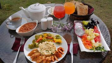 A round glass table with a meal consisting of vegetables, chicken, and a fruit platter, along with drinks and condiments.