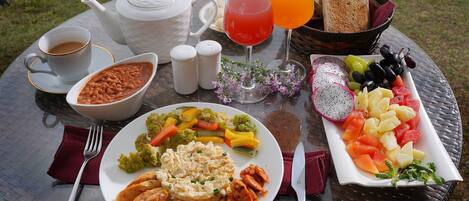 A round glass table with a meal consisting of vegetables, chicken, and a fruit platter, along with drinks and condiments.