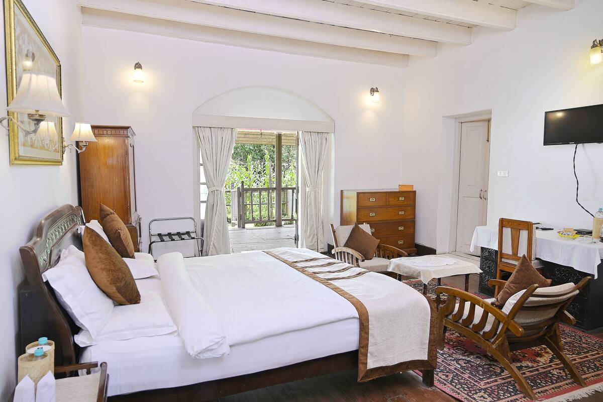 A hotel room with a large bed, two chairs, a wooden dresser, a television, and a balcony with curtains.