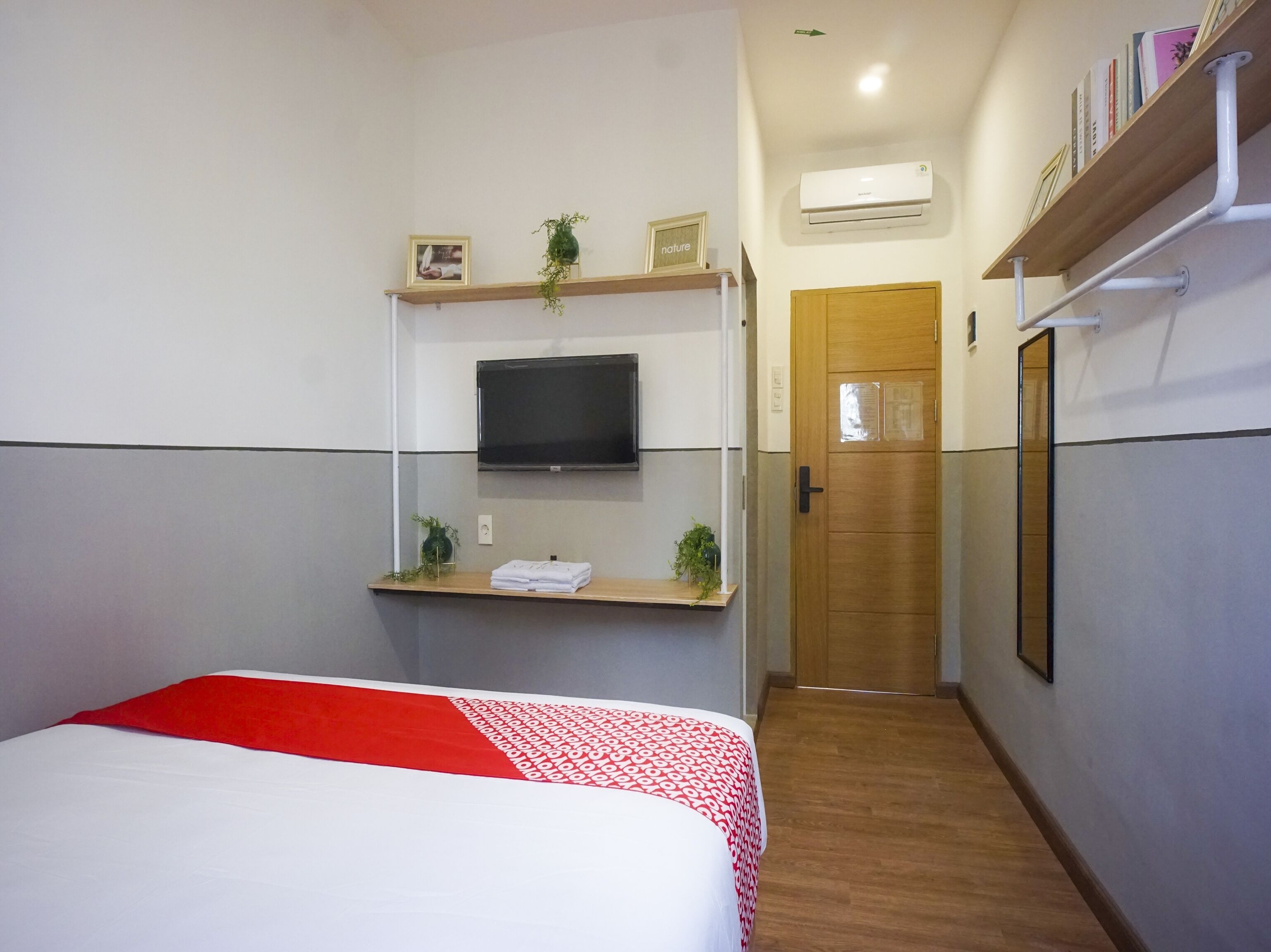 A hotel room with a bed, a TV mounted on a wall, a wooden door, and a shelf with books and a plant.