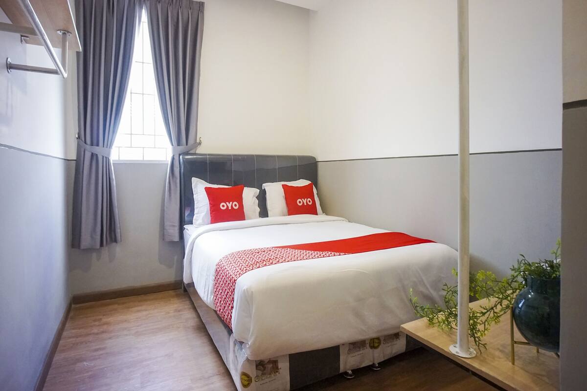 A neatly arranged hotel room with a bed, a window with curtains, a bedside table with a plant, and a wall-mounted shelf.
