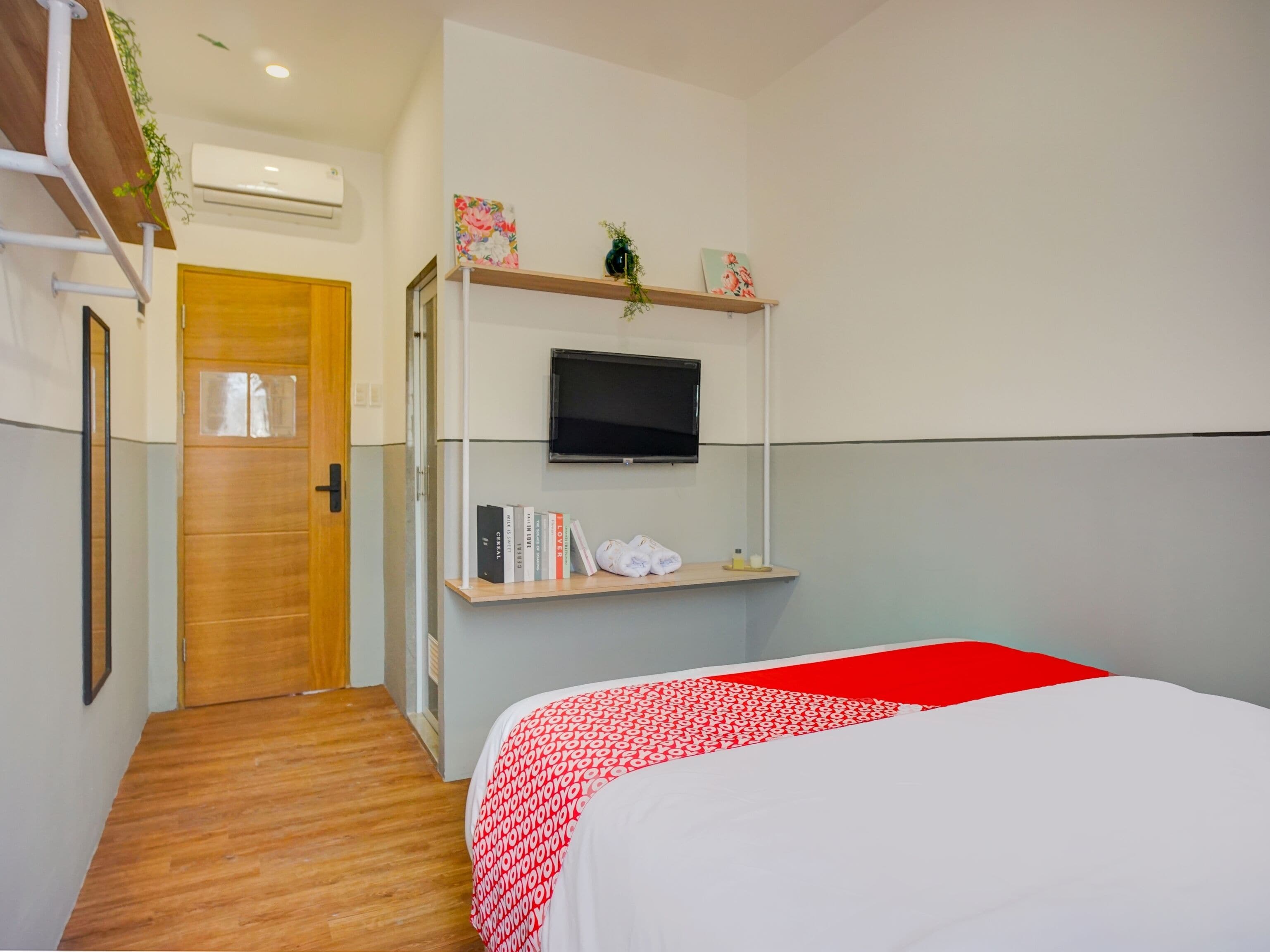 Ett hotellrum med en dörr av trä, en säng med ett rött och vitt mönstrat överkast, en väggmonterad tv, en hylla med böcker och en växt, samt ett trägolv.