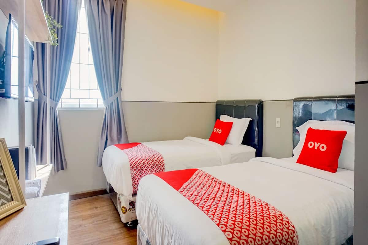 A hotel room with two beds, each with a red pillow and a white bedspread. There is a window with curtains, a framed picture on the left, and a small table with a remote control.