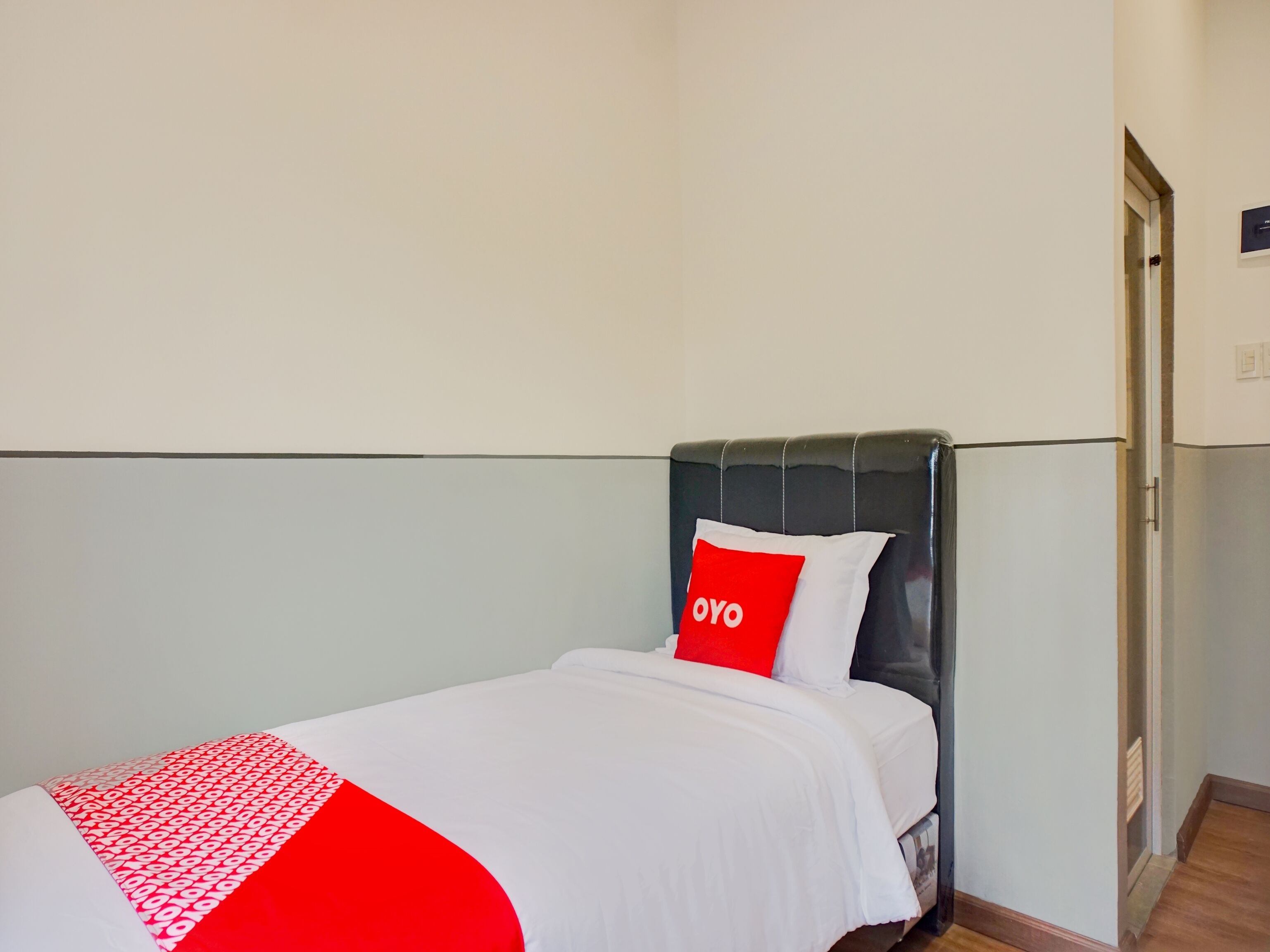 Una cama individual con cabecera negra, ropa de cama blanca y una almohada roja con la palabra 'OYO' escrita.