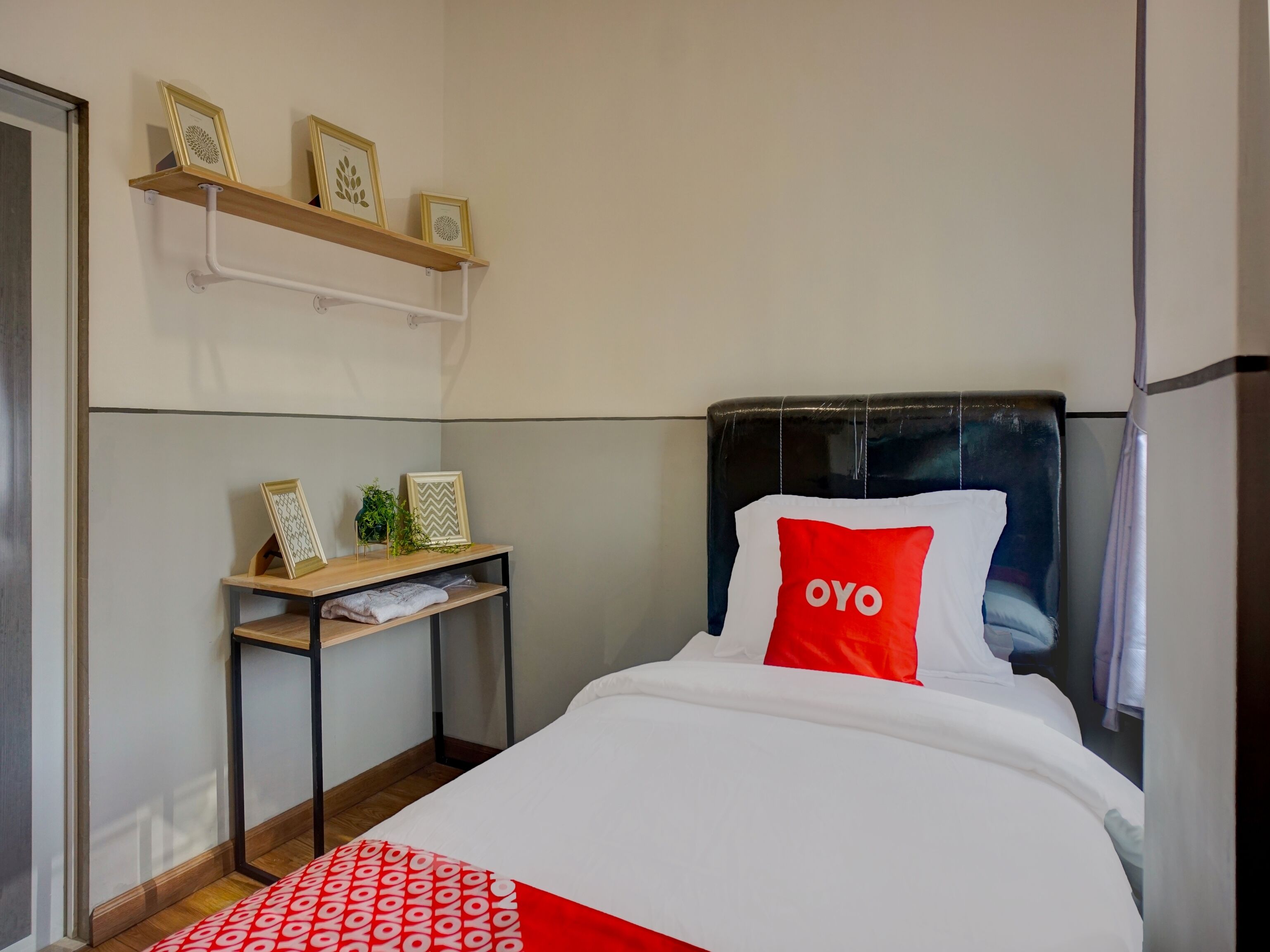 A neatly arranged hotel room with a bed, a bedside table with a plant, and a shelf with framed pictures.