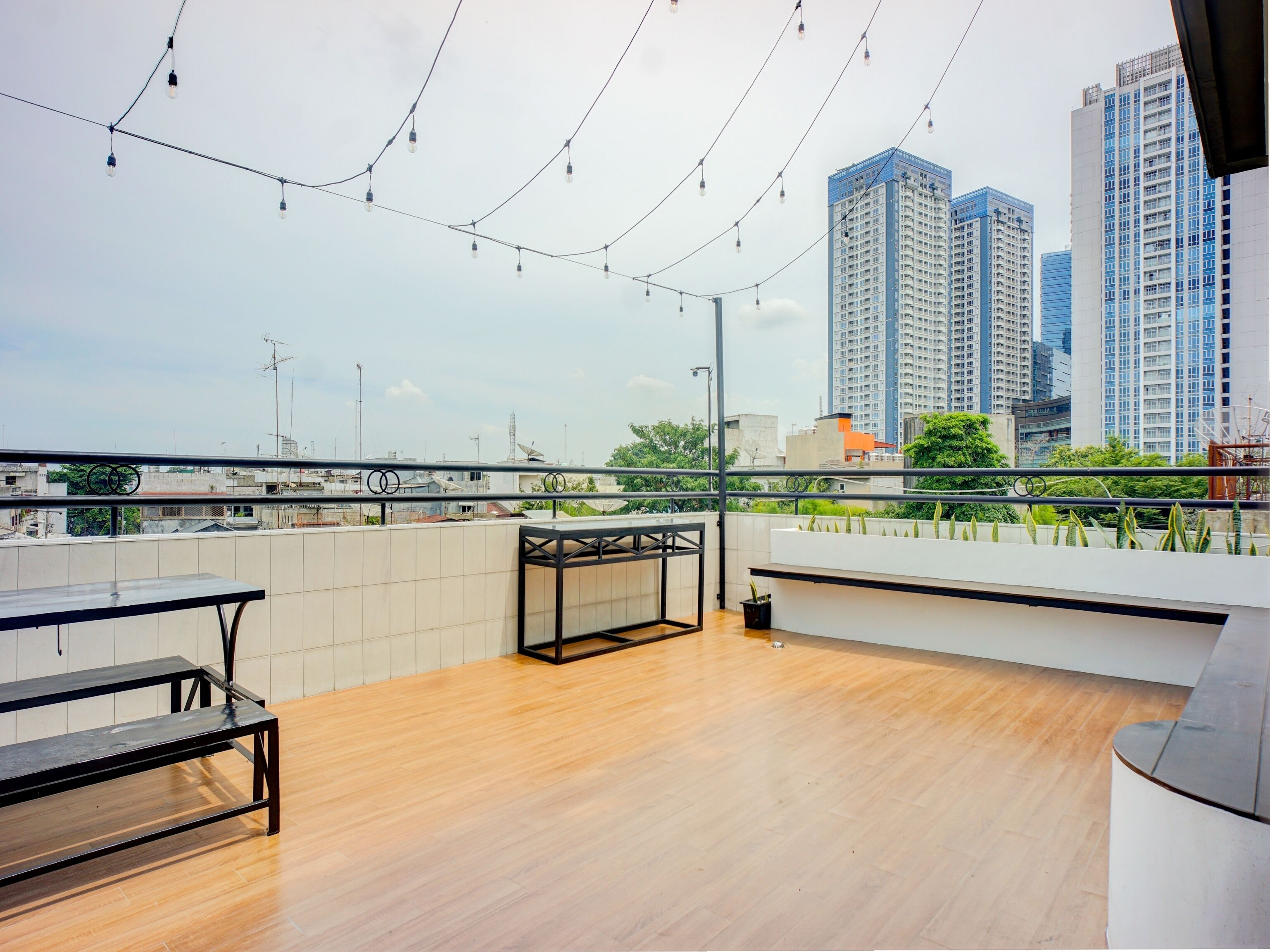 Eine Dachterrasse mit Holzboden, einem schwarzen Metalltisch und Lichterketten. Im Hintergrund Hochhäuser und ein klarer Himmel.