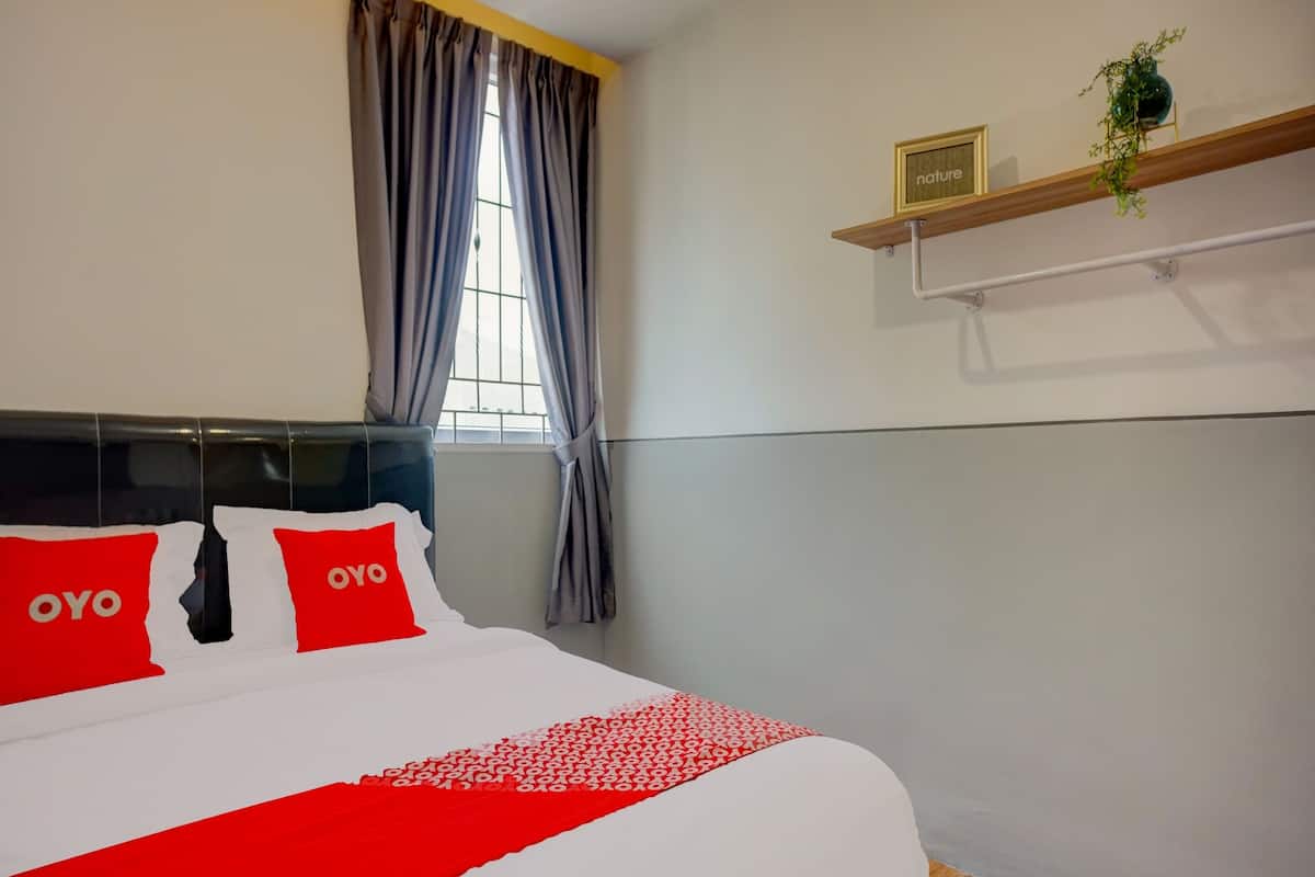 A hotel room with a bed featuring red and white bedding, a window with grey curtains, and a shelf with a plant and a framed picture.