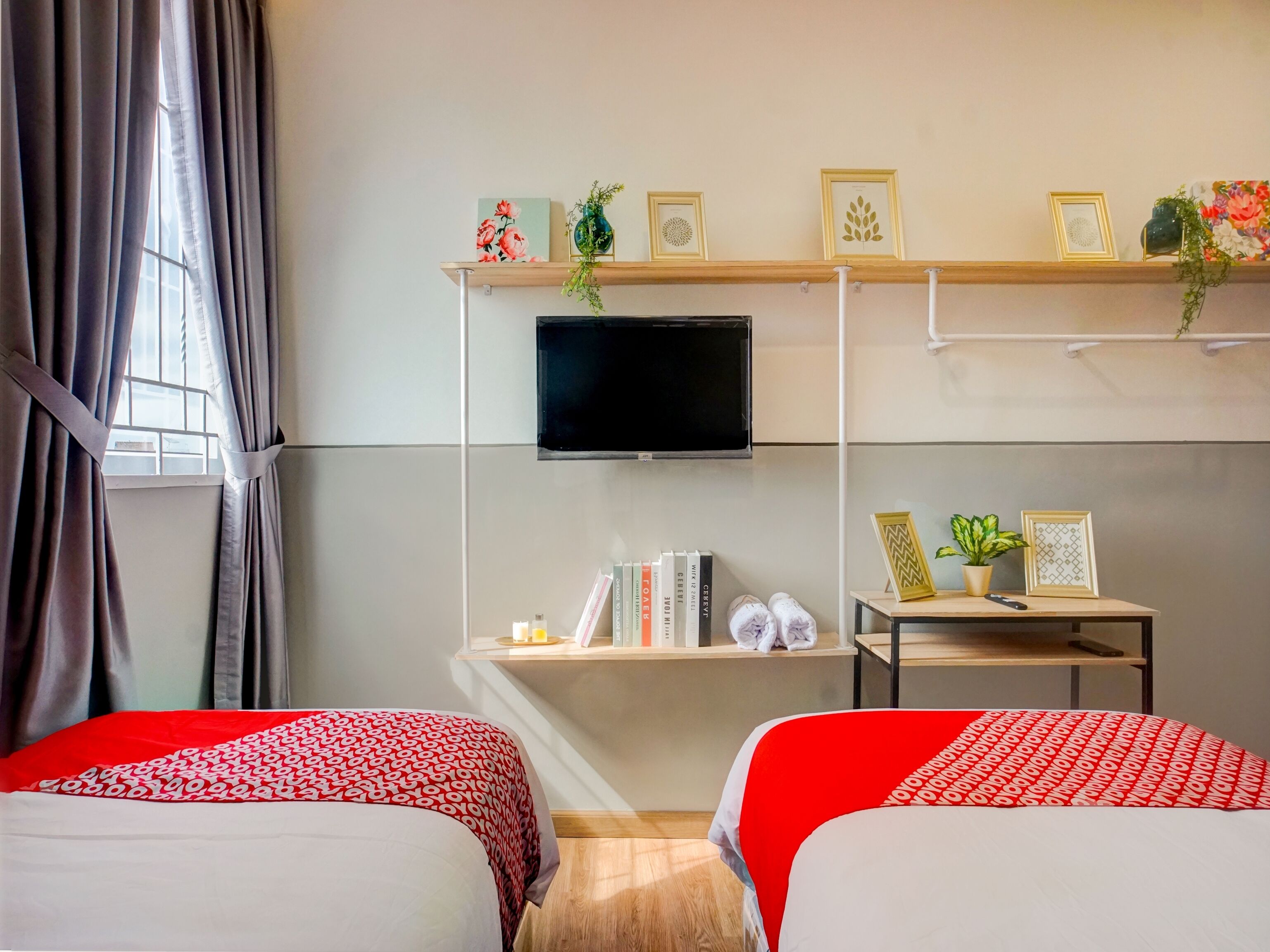 A hotel room with two beds, a TV, a shelf with books and decorative items, and a window with curtains.