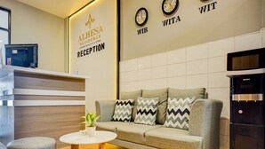 A reception area with a reception desk, a grey sofa with patterned cushions, a small round table with a plant, and a wall clock labeled 'WITA'. - Hotel O Alhesa Residence (Medan)