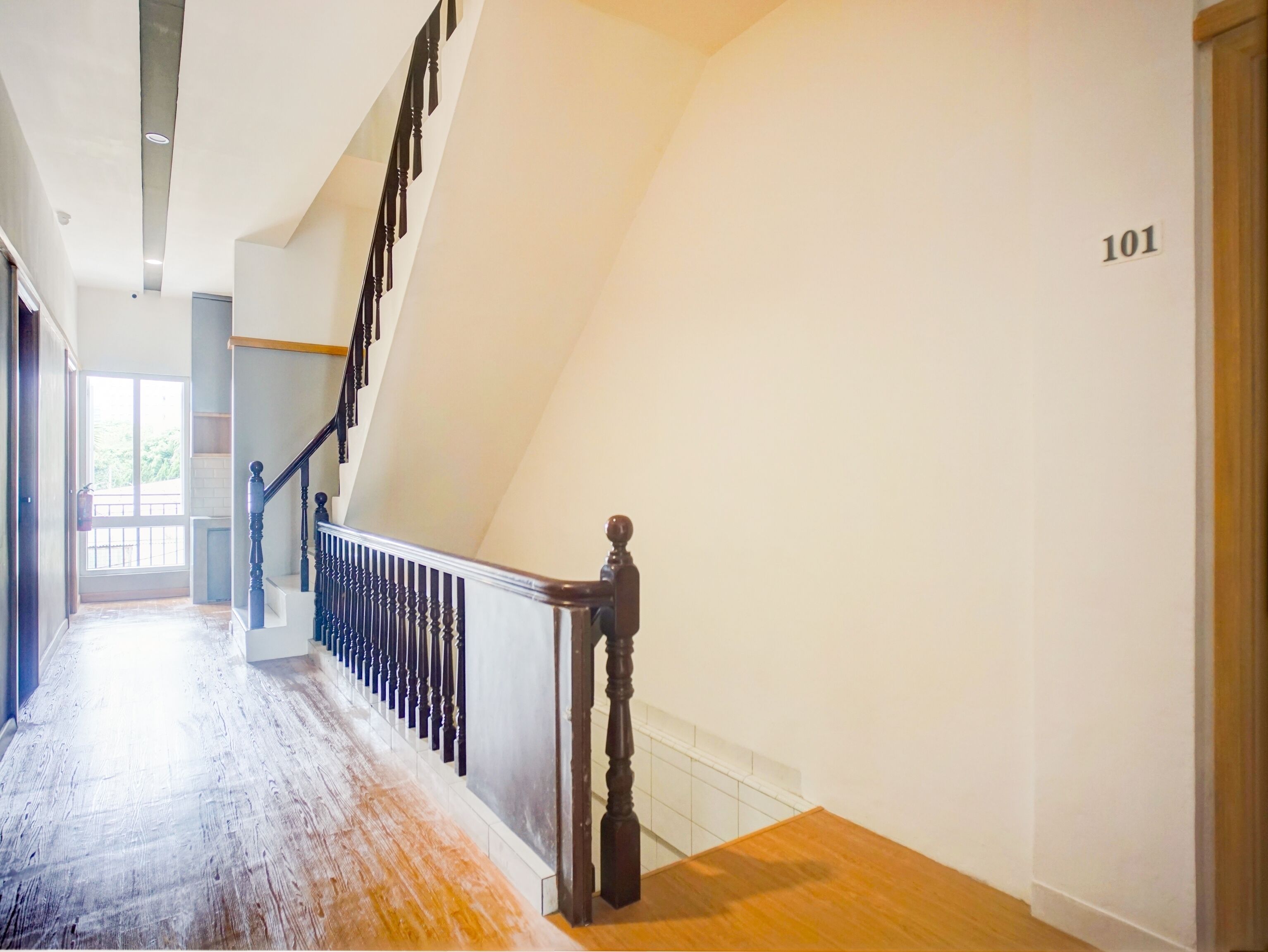 Un pasillo espacioso con piso de madera, una escalera con barandales oscuros y una pared blanca con el número 101.