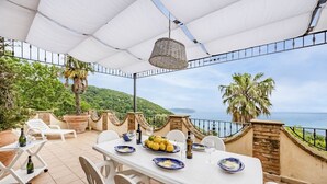 An outdoor patio with a table set for dining, overlooking a scenic view of the sea and hills.