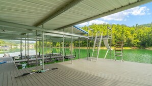 Une terrasse en bois couverte, dotée de tables de pique-nique et d’une échelle menant à un quai surplombant un lac paisible, le tout entouré d’arbres.