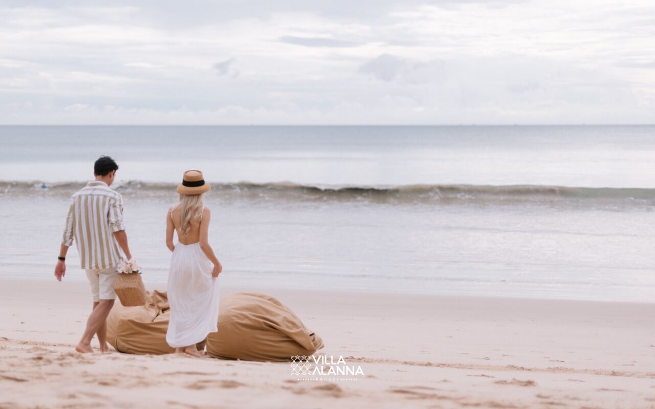 Um casal na praia, com um saco de areia, uma camisa listrada e uma saia branca.