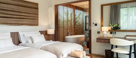 A hotel room with two beds, a sitting area with a chair, a desk with a lamp, a large mirror, and a bathroom with a bathtub.